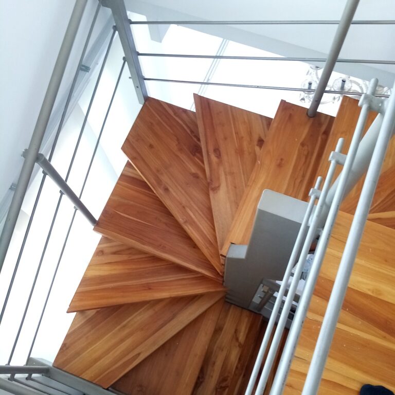 Vista desde la parte alta hacia abajo de una escalera de caracol con peldaños de madera y pasamanos de tubos paralelos de acero.