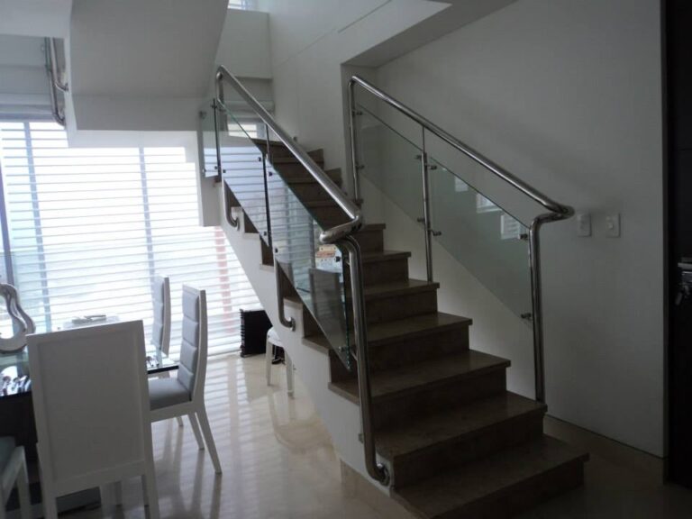 Vista interior de una escalera residencial con peldaños de madera y una elegante baranda compuesta por pasamanos de acero inoxidable y paneles de vidrio templado transparente.
