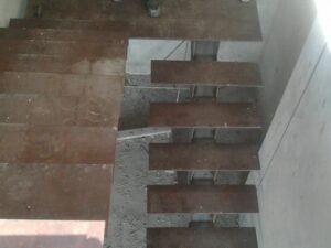Vista superior de una escalera en construcción con peldaños de chapa de acero sin pintar sobre una base de concreto y paredes sin revestir.