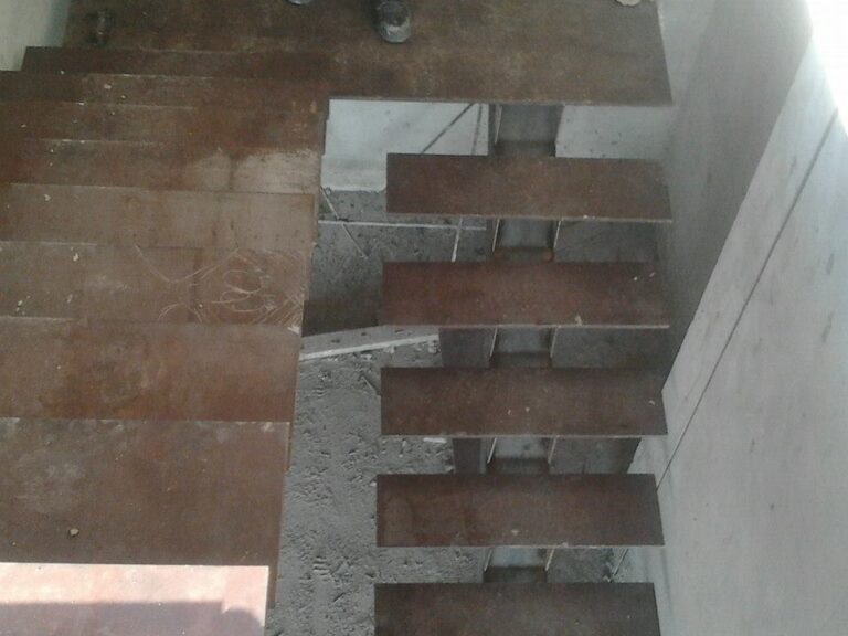 Vista superior de una escalera en construcción con peldaños de chapa de acero sin pintar sobre una base de concreto y paredes sin revestir.
