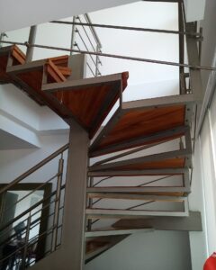 Perspectiva ascendente de una escalera de caracol de estructura metálica gris y peldaños triangulares de madera barnizada, con baranda de tubos metálicos.