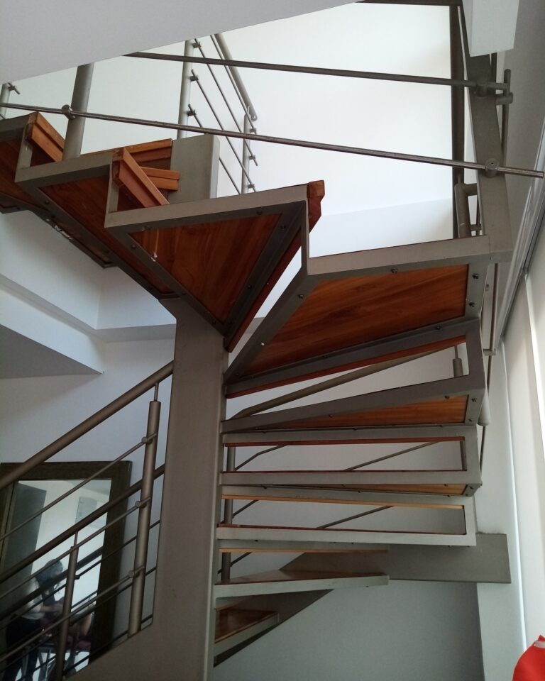 Perspectiva ascendente de una escalera de caracol de estructura metálica gris y peldaños triangulares de madera barnizada, con baranda de tubos metálicos.
