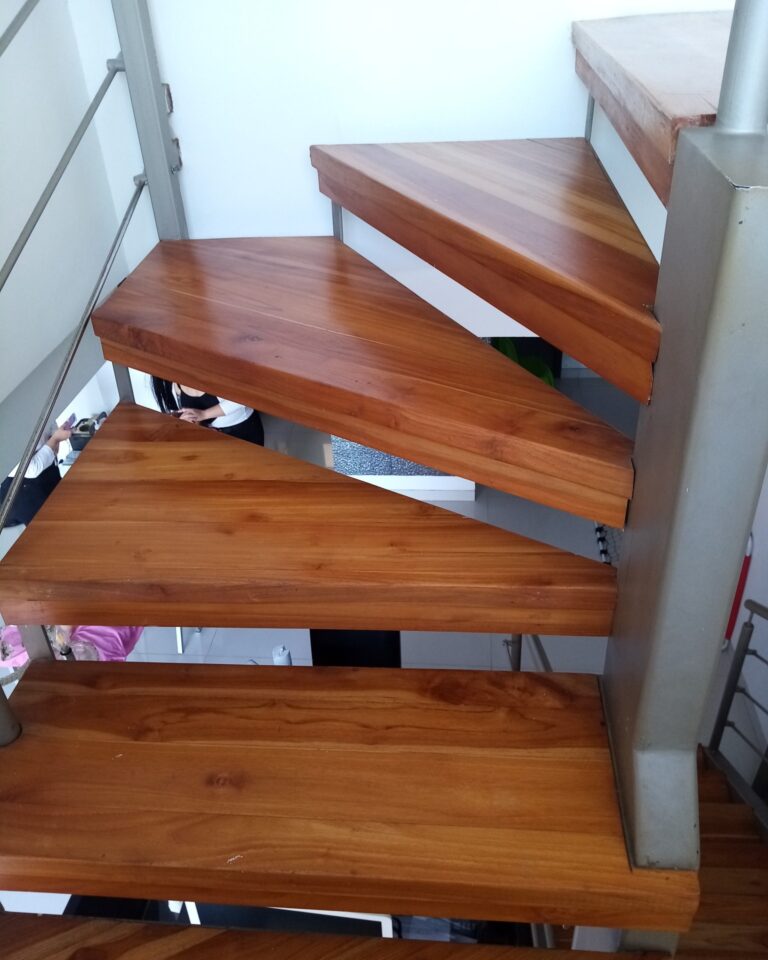 Primer plano de peldaños triangulares de madera barnizada brillante en una escalera de caracol con columna de soporte metálica.