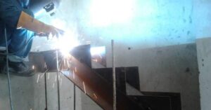 Un trabajador con equipo de protección realiza una soldadura eléctrica en una viga metálica en zigzag para una escalera de obra.