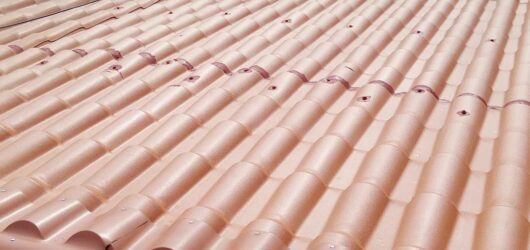 Vista en primer plano de un techo inclinado cubierto con tejas metálicas esmaltadas de color terracota. Las tejas imitan la forma tradicional de teja española y están fijadas con tornillos visibles en cada onda. Al fondo se ve una parte de un techo antiguo con tejas de asbesto rojas deterioradas.