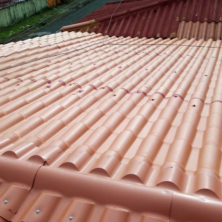 Vista en primer plano de un techo inclinado cubierto con tejas metálicas esmaltadas de color terracota. Las tejas imitan la forma tradicional de teja española y están fijadas con tornillos visibles en cada onda. Al fondo se ve una parte de un techo antiguo con tejas de asbesto rojas deterioradas.