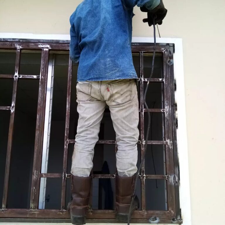 Un herrero, de espaldas a la cámara, está de pie sobre una base improvisada para alcanzar la parte superior de una reja de ventana de metal marrón, de la cual está pintando el marco con una brocha. Viste una camisa de mezclilla azul y pantalones de mezclilla beige. En la parte superior derecha, su mano enguantada sostiene un objeto. Paredes de color crema detrás.