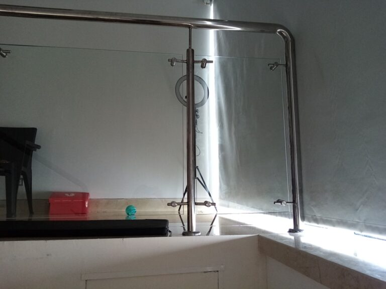Vista cercana de una barandilla para balcón o escalera hecha con tubos y postes de acero inoxidable que sujetan paneles de vidrio transparente, con una pared blanca y una luz de aro al fondo.