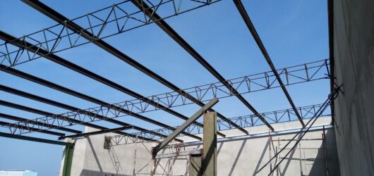 Vista desde el interior de una obra en construcción, mirando hacia arriba a una estructura de techo abierta formada por vigas de acero y cerchas de celosía. Debajo se aprecian paredes de concreto, andamios metálicos y un cielo azul despejado al fondo.