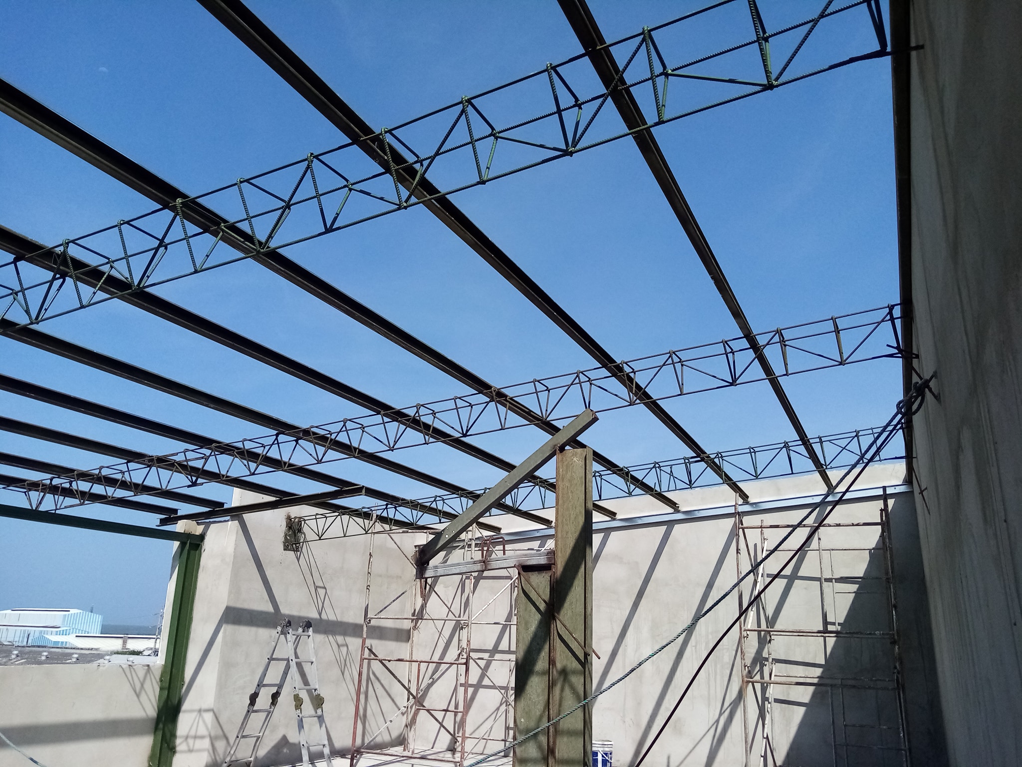 Vista desde el interior de una obra en construcción, mirando hacia arriba a una estructura de techo abierta formada por vigas de acero y cerchas de celosía. Debajo se aprecian paredes de concreto, andamios metálicos y un cielo azul despejado al fondo.