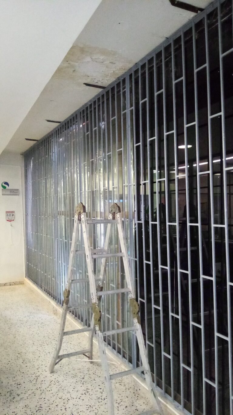 Una escalera de mano de aluminio está colocada frente a una gran ventana de oficina cubierta por una reja de seguridad de metal de barrotes verticales grises. El piso es de terrazo y las paredes blancas. Letreros de seguridad son visibles en la pared izquierda.